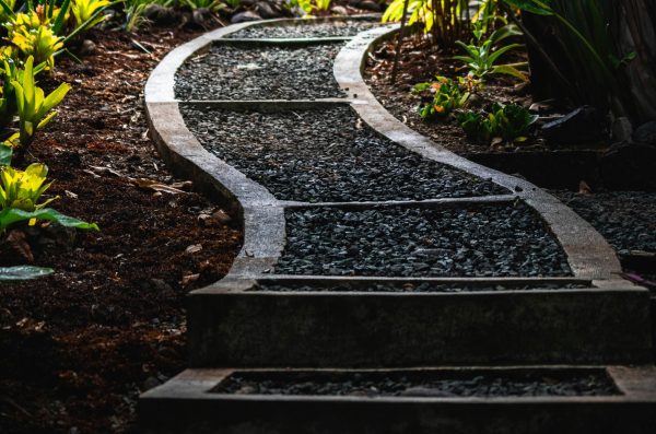 A winding path with contrasting dark and light stones, cutting through a lush sloped garden filled with vibrant foliage.