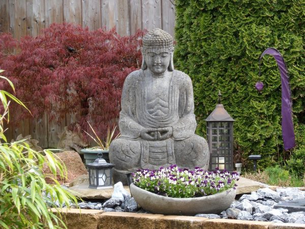 Serene garden featuring a stone Buddha statue, surrounded by vibrant red maple, greenery, and a bowl of purple flowers.